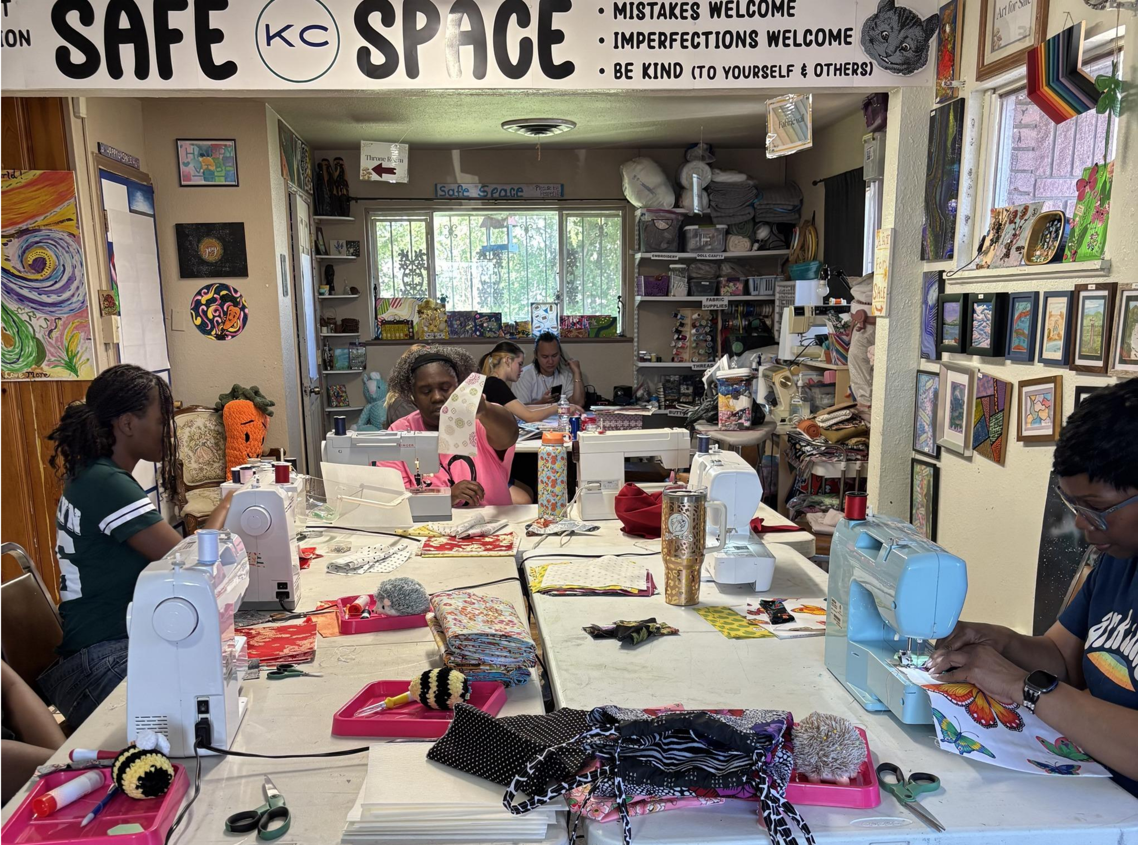 Sewing class with diverse adults focused on projects in a cozy, art-filled room. A Safe Space sign promotes kindness and embracing imperfections.