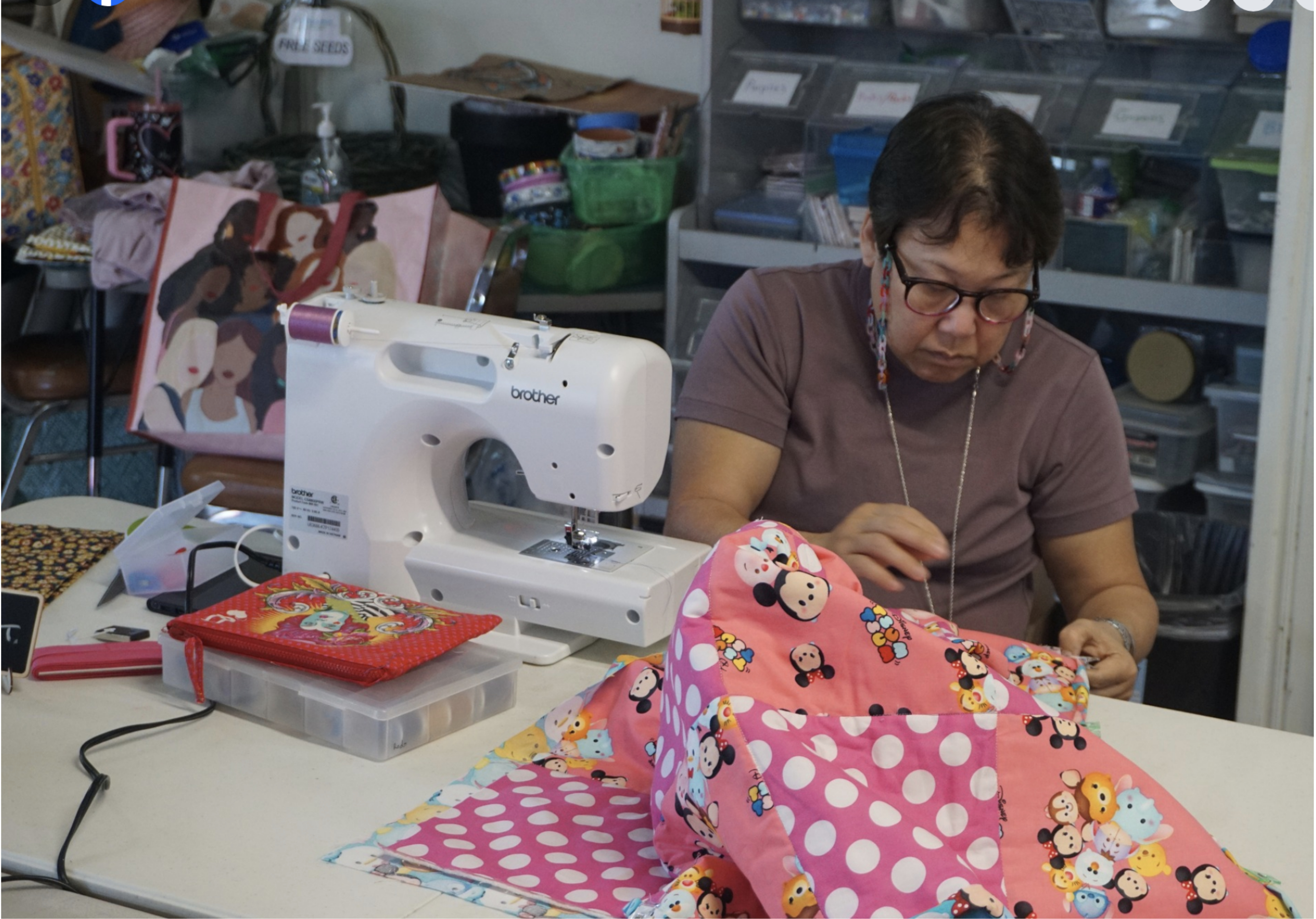 A person sewing at a table with a white sewing machine, working on a colorful fabric featuring cartoon characters. The workspace is filled with organized supplies.