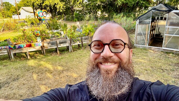 New AmeriCorps member Drew Nevitt in the garden he tends, with raised beds and the greenhouse behind him.
