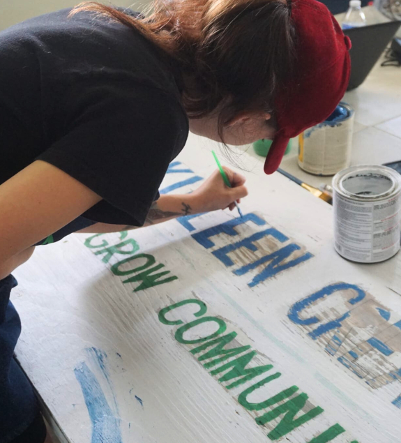 A women painting a sign with the green and blue Killeen Creators logo on to a wooden surface. Various paint supplies are visible on the table.